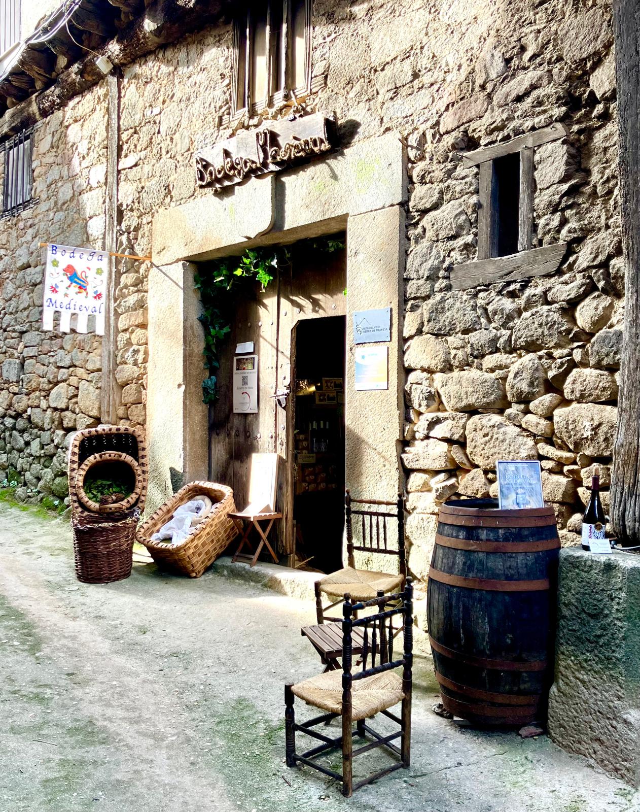 Entrada de piedra con letrero “Bodega Medieval”, barril de madera, sillas y cestas junto a una puerta abierta.
